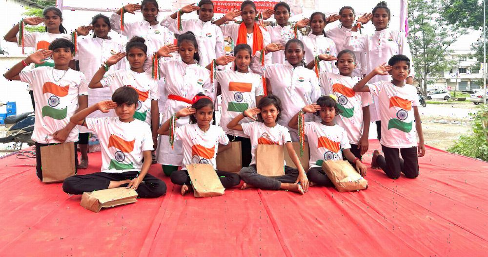 Children in white T-shirts saluting during a national day celebration.