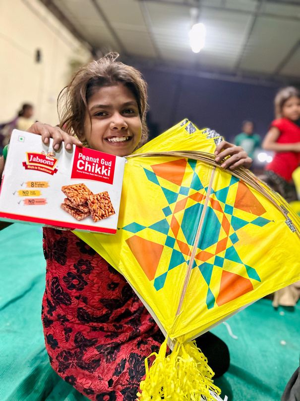 A girl smiling while holding a box of chikki and a colourful kite.